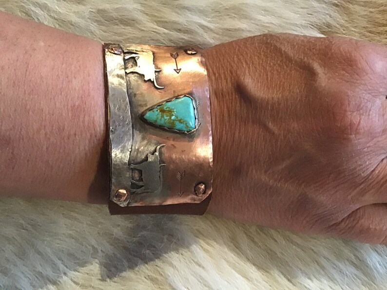 May include: A copper cuff bracelet with a turquoise stone and silver accents. The bracelet features a design of two stylized cattle and arrows.