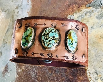 May include: A brown leather cuff bracelet with a copper plate featuring three turquoise stones and etched designs of suns and arrows.