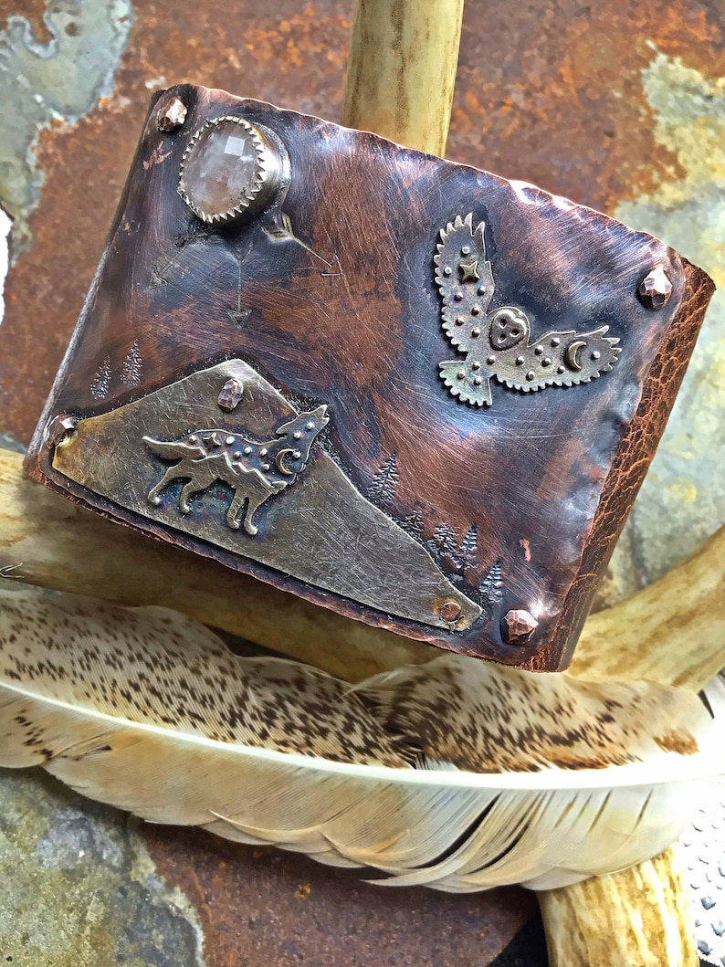 Puede incluir: Un brazalete de cobre con un lobo aullando a la luna, un b&uacute;ho volando con estrellas y una piedra de cuarzo transparente. El brazalete est&aacute; decorado con remaches de cobre y tiene un acabado martillado.