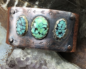 May include: A copper cuff bracelet with three turquoise stones set in silver. The bracelet is decorated with arrows and suns.