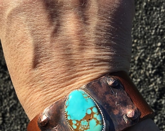 May include: A wide cuff bracelet featuring a large, oval turquoise stone set in a silver bezel. The copper band has decorative arrow and dot designs. The bracelet is attached to a brown leather strap.