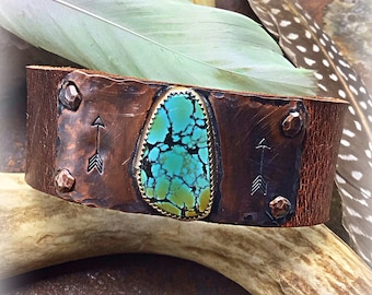 May include: A brown leather cuff bracelet with a turquoise stone set in a copper frame. The bracelet has two arrows engraved on the copper. The bracelet is accented with copper rivets.