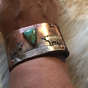 May include: A copper and silver cuff bracelet with a turquoise stone and two steer silhouettes. The bracelet is on a brown leather band.