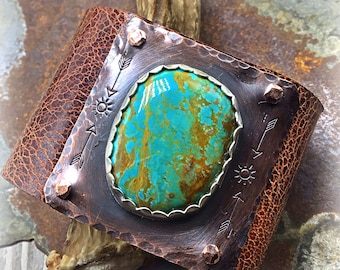 May include: A copper and leather cuff bracelet with a turquoise stone inlay. The stone is set in a silver bezel and the cuff is decorated with arrows and suns.