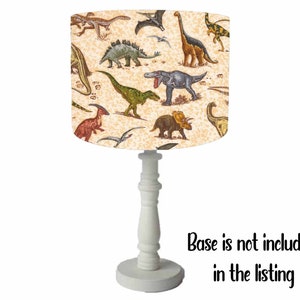May include: A white table lamp with a cream-colored drum shade featuring a dinosaur print. The shade has a variety of dinosaurs in different colors and poses. The base is white and has a classic design. Base is not included in the listing.