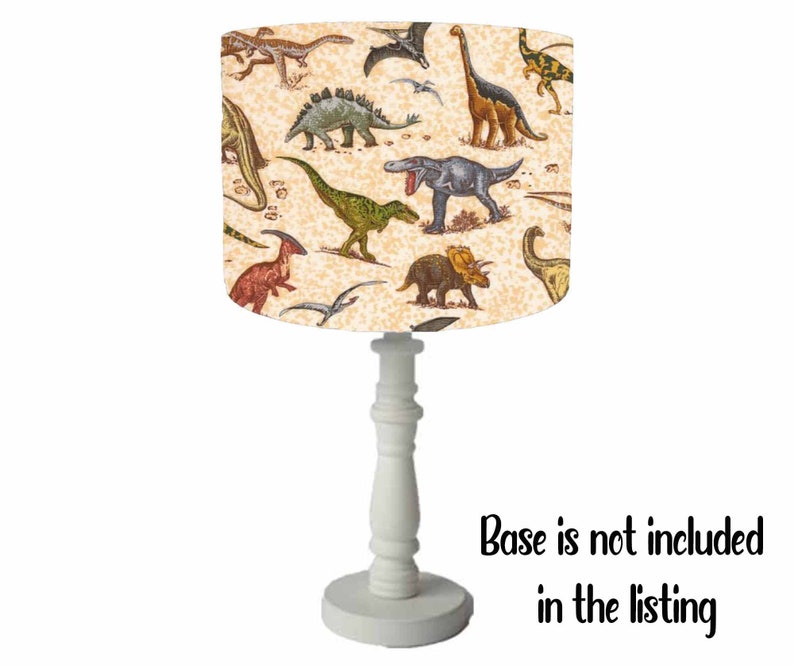 May include: A white table lamp with a cream-colored drum shade featuring a dinosaur print. The shade has a variety of dinosaurs in different colors and poses. The base is white and has a classic design. Base is not included in the listing.