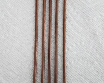 May include: Five wooden knitting needles, all the same size and color, are arranged in a row on a white surface. The needles are brown and have a pointed tip.