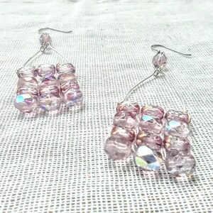 Puede incluir: Un par de pendientes colgantes con un marco de alambre plateado y múltiples cuentas de vidrio rosa iridiscentes.