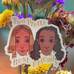 May include: A sticker featuring two illustrated faces of diverse women with the text "Two Pretty Best Friends" in a peach color. The sticker is surrounded by dried flowers in yellow, purple, and brown.