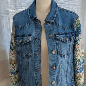May include: A denim jacket with crochet sleeves. The jacket has a classic design with two chest pockets and button closures. The sleeves are made of colorful crochet yarn in shades of blue, pink, green, and yellow.