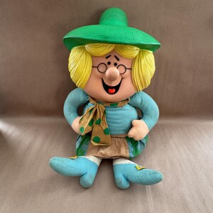May include: A whimsical, vintage wall hanging featuring a smiling, cartoon-like figure. The figure has blonde hair, glasses, and a green hat. It wears a blue shirt, a patterned scarf, and blue shoes. The overall design is colorful and cheerful.