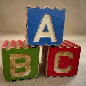 以下が含まれることがあります： 積み重ねられた3つのヴィンテージ木製アルファベットブロック。一番上のブロックは青色で白い「A」、左のブロックは緑色で白い「B」、右のブロックは赤色で白い「C」です。ブロックは使い古された外観をしています。