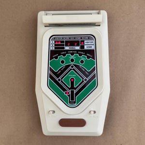 May include: A vintage baseball game with a white plastic casing. The game features a baseball field graphic in green and black, with scoring displays for visitor and home teams. The game has buttons for on/off and auto/man.