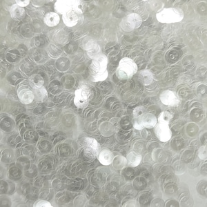 Puede incluir: Primer plano de un montón de lentejuelas blancas iridiscentes. Las lentejuelas son redondas y tienen un centro ligeramente elevado.