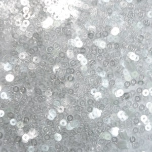 Puede incluir: Primer plano de un montón de lentejuelas plateadas. Las lentejuelas son pequeñas y redondas, y están esparcidas al azar.