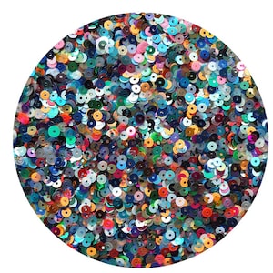 May include: A circular arrangement of colorful sequins in various sizes and shades. The sequins are primarily in shades of blue, green, silver, and red, creating a shimmering, textured effect. The sequins are scattered across a white background.