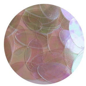 May include: A close-up view of a collection of iridescent, oval-shaped sequins. The sequins are a mix of pale pink, green, and clear, overlapping each other in a circular pattern against a white background. The sequins have a subtle shimmer.