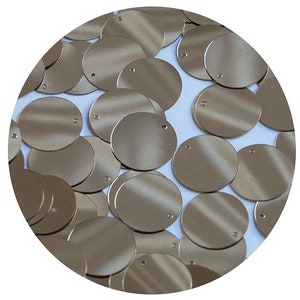 May include: A collection of metallic gold-colored sequins, each with a small hole at the top for stringing. The sequins are round and have a matte finish. They are scattered across a white surface.