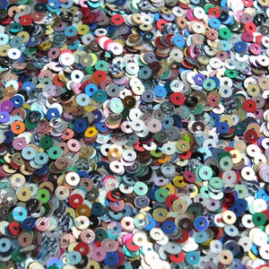 May include: A close-up view of a pile of colorful sequins. The sequins are round with a hole in the center, and they come in a variety of colors, including red, blue, green, silver, and gold. The sequins are scattered and overlapping.