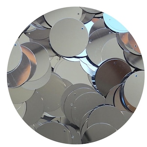 May include: A close-up of a collection of silver, circular sequins. Each sequin has a small hole near the edge. The sequins are scattered, overlapping, and reflecting light, creating a shiny, metallic appearance. The background is white.