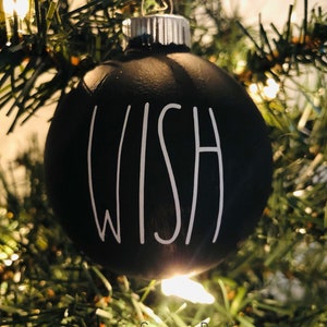 May include: A black Christmas ornament with the word "WISH" written in white on it. The ornament is hanging on a green Christmas tree.