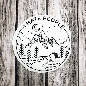 May include: A white sticker with a black line drawing of a mountain scene. The design includes mountains, trees, a crescent moon, stars, and a tent. The text "I HATE PEOPLE" is written around the top edge of the sticker.
