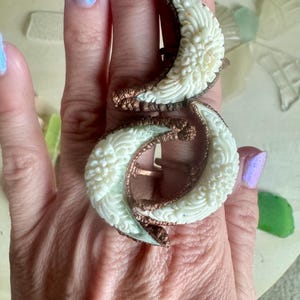 May include: An ornate ring featuring two cream-colored, crescent-shaped carvings with floral details. The carvings are connected by a copper-colored metal band. The ring is worn on a finger, showcasing its intricate design and craftsmanship.