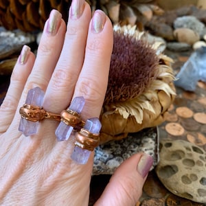 May include: A unique three-stone ring featuring three light purple crystal points set in copper-colored metal. The ring is worn on a hand with iridescent nail polish. The background includes natural elements like pinecones and dried flowers.