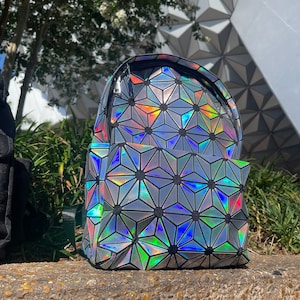 May include: A small iridescent silver backpack with a geometric pattern. The backpack has a black zipper and a black strap.