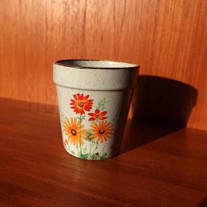 Può includere: Un piccolo vaso di fiori in ceramica bianco sporco con finitura maculata e bordo marrone. Il vaso è decorato con disegni floreali arancioni e rossi. Il vaso è appoggiato su una superficie di legno.