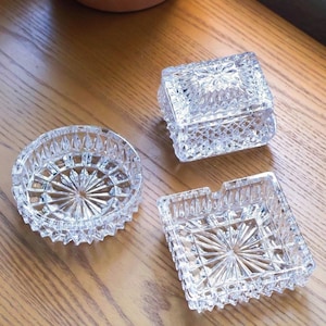 May include: A set of three clear glass items featuring intricate geometric cut patterns. The collection includes a lidded rectangular box, a circular dish with a starburst base, and a square tray with a notched edge and a starburst center.