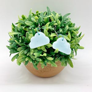 May include: A pair of light blue cloud-shaped earrings with a smooth, glossy finish. Each earring features a silver-toned metal hook and a small light blue jump ring connecting the cloud charm to the ear wire, creating a simple dangling design.