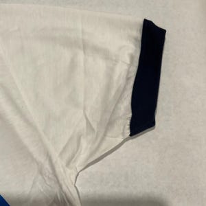 Puede incluir: Camiseta blanca con pu&ntilde;o de manga azul marino. La camiseta tiene un dise&ntilde;o sencillo y parece estar hecha de una tela suave y ligera. El pu&ntilde;o de la manga es de color azul marino liso.