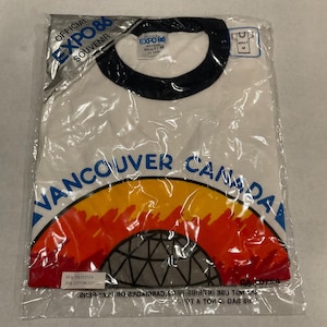 Puede incluir: Camiseta blanca en una bolsa de pl&aacute;stico con cuello negro. La camiseta presenta el texto "VANCOUVER CANADA" sobre un gr&aacute;fico colorido y el texto "EXPO 86 OFFICIAL SOUVENIR". La camiseta es talla Adulto/M.