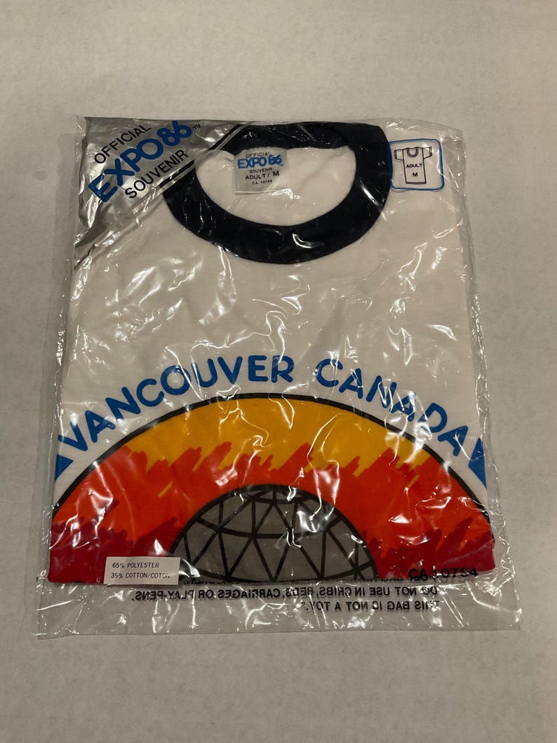Puede incluir: Camiseta blanca en una bolsa de pl&aacute;stico con cuello negro. La camiseta presenta el texto "VANCOUVER CANADA" sobre un gr&aacute;fico colorido y el texto "EXPO 86 OFFICIAL SOUVENIR". La camiseta es talla Adulto/M.