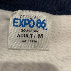 Puede incluir: Primer plano de una etiqueta de camiseta blanca con el texto "OFFICIAL EXPO 86 SOUVENIR ADULT/M CA 10754". La camiseta tiene cuello y ribetes de manga azul marino.