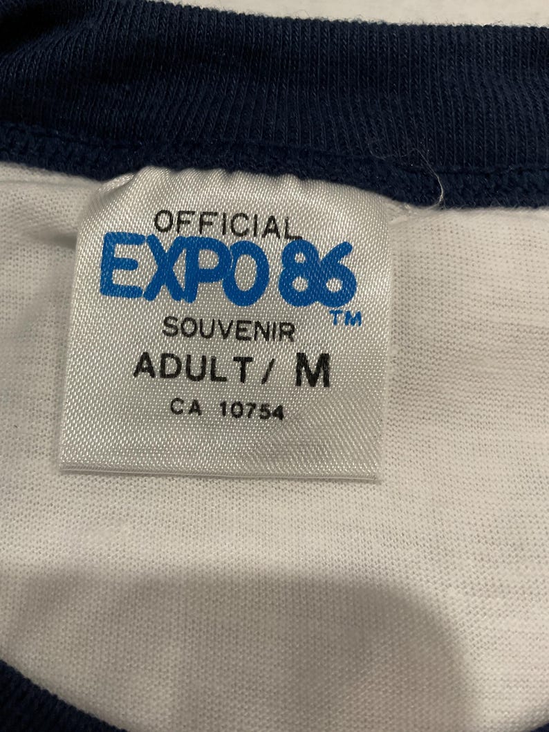 Puede incluir: Primer plano de una etiqueta de camiseta blanca con el texto "OFFICIAL EXPO 86 SOUVENIR ADULT/M CA 10754". La camiseta tiene cuello y ribetes de manga azul marino.