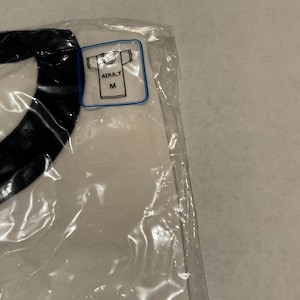 Puede incluir: Una camiseta blanca con un dise&ntilde;o negro, sellada en una bolsa de pl&aacute;stico transparente. Una peque&ntilde;a etiqueta indica que la camiseta es de talla mediana para adultos.