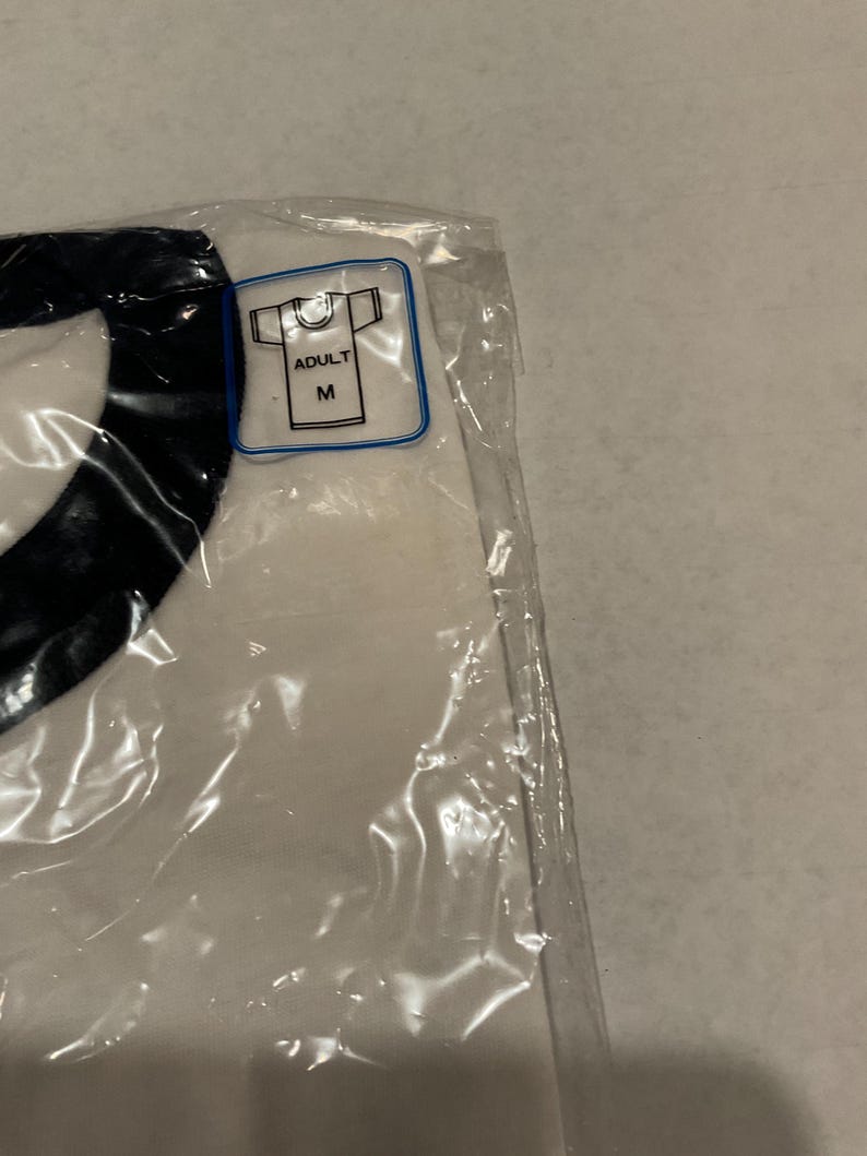 Puede incluir: Una camiseta blanca con un dise&ntilde;o negro, sellada en una bolsa de pl&aacute;stico transparente. Una peque&ntilde;a etiqueta indica que la camiseta es de talla mediana para adultos.