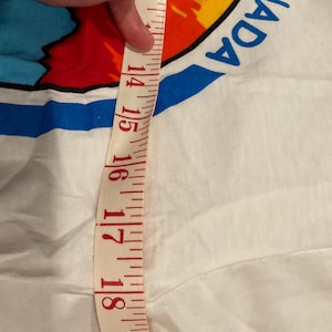 Puede incluir: Camiseta blanca con un gr&aacute;fico colorido y la palabra "CANADA" en azul. Una cinta m&eacute;trica muestra una longitud de aproximadamente 35,5 a 45,7 cm. La cinta es roja y blanca.