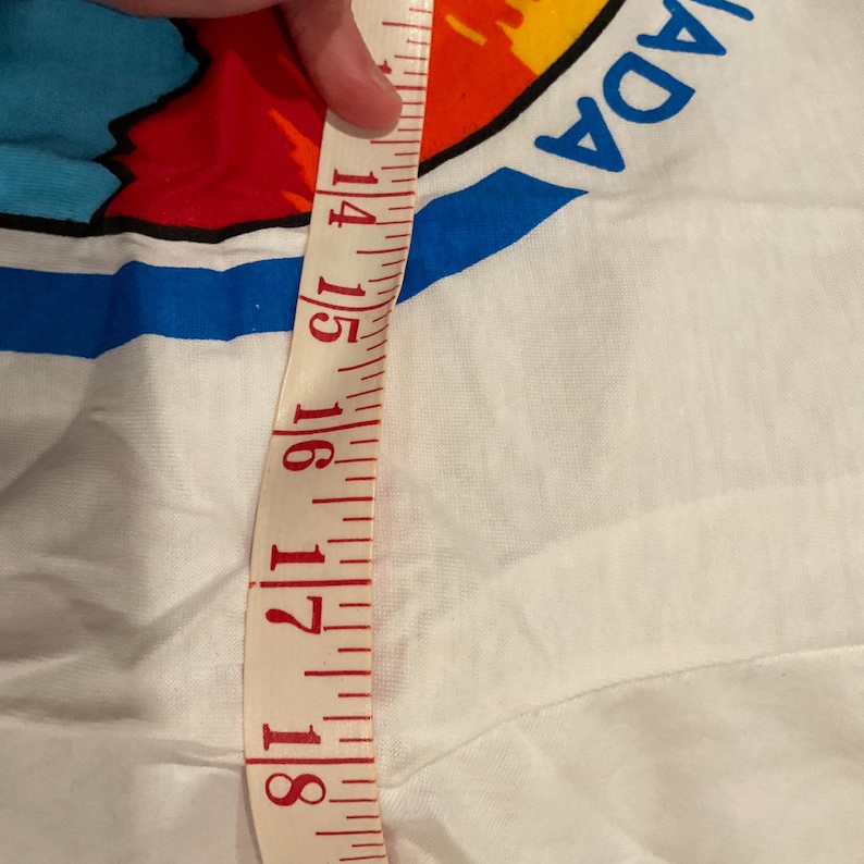 Puede incluir: Camiseta blanca con un gr&aacute;fico colorido y la palabra "CANADA" en azul. Una cinta m&eacute;trica muestra una longitud de aproximadamente 35,5 a 45,7 cm. La cinta es roja y blanca.