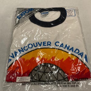 Puede incluir: Una camiseta blanca con cuello redondo, a&uacute;n en su embalaje original, con el texto "VANCOUVER CANADA" en azul, y un gr&aacute;fico colorido de una c&uacute;pula y un arco&iacute;ris. La camiseta es un recuerdo oficial de la Expo 86.