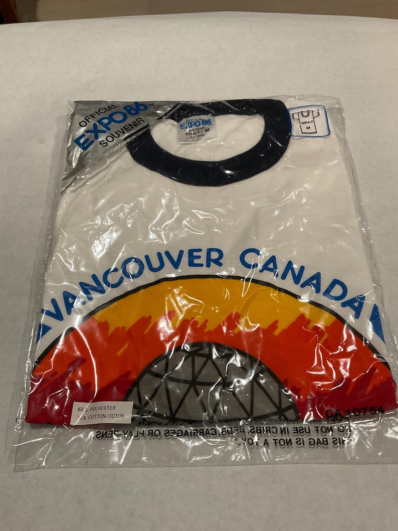 Puede incluir: Una camiseta blanca con cuello redondo, a&uacute;n en su embalaje original, con el texto "VANCOUVER CANADA" en azul, y un gr&aacute;fico colorido de una c&uacute;pula y un arco&iacute;ris. La camiseta es un recuerdo oficial de la Expo 86.
