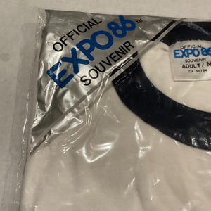 Puede incluir: Una camiseta blanca con cuello azul marino, empaquetada en una bolsa de pl&aacute;stico transparente. La camiseta presenta el texto "OFFICIAL EXPO 86 SOUVENIR" en azul. La etiqueta dice "ADULT/M".