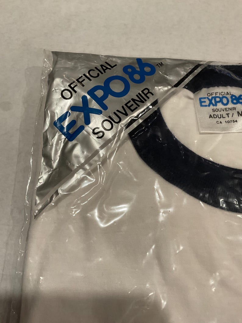 Puede incluir: Una camiseta blanca con cuello azul marino, empaquetada en una bolsa de pl&aacute;stico transparente. La camiseta presenta el texto "OFFICIAL EXPO 86 SOUVENIR" en azul. La etiqueta dice "ADULT/M".
