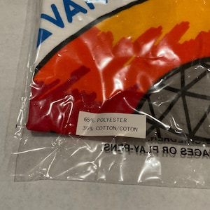 Puede incluir: Una toalla blanca en una bolsa de pl&aacute;stico con la palabra "VANCOUVER" en azul. La toalla tiene un dise&ntilde;o colorido con tonos naranja, amarillo y rojo. La etiqueta dice "65% POLYESTER 35% COTTON/COTON."