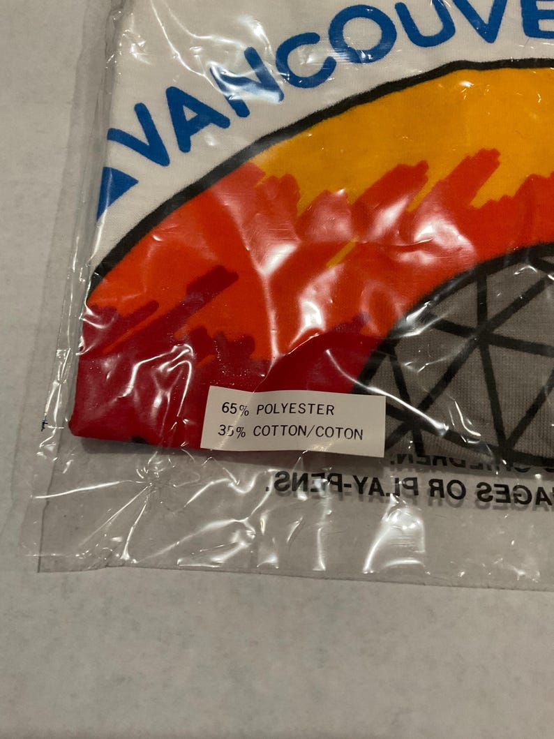 Puede incluir: Una toalla blanca en una bolsa de pl&aacute;stico con la palabra "VANCOUVER" en azul. La toalla tiene un dise&ntilde;o colorido con tonos naranja, amarillo y rojo. La etiqueta dice "65% POLYESTER 35% COTTON/COTON."