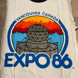 Puede incluir: Camiseta blanca con cuello y ribetes de manga azul marino. La parte delantera presenta un gr&aacute;fico colorido de una estructura similar a un globo con el texto "VANCOUVER CANADA" y "EXPO 86" en azul.