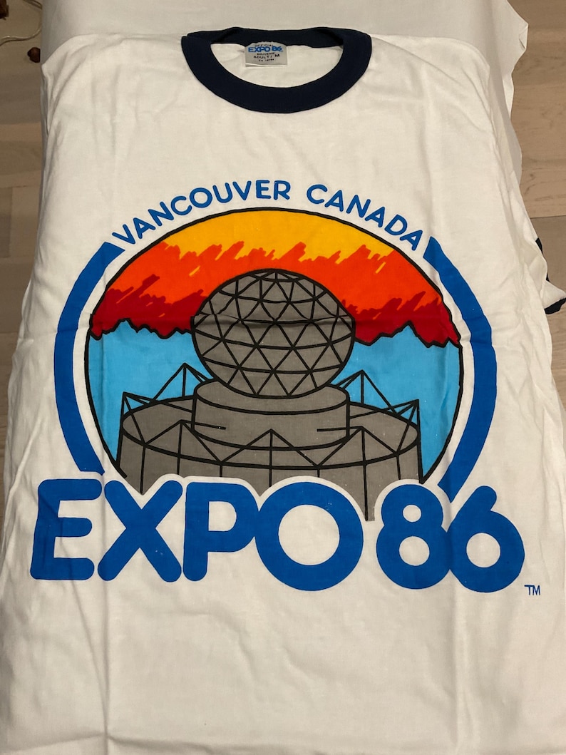 Puede incluir: Camiseta blanca con cuello y ribetes de manga azul marino. La parte delantera presenta un gr&aacute;fico colorido de una estructura similar a un globo con el texto "VANCOUVER CANADA" y "EXPO 86" en azul.