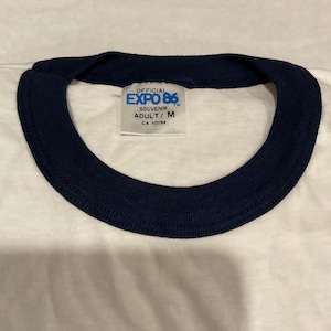 Puede incluir: Camiseta blanca con cuello azul marino. La camiseta presenta el texto "OFFICIAL EXPO 86" en una etiqueta. La etiqueta tambi&eacute;n incluye el texto "SOUVENIR ADULT/ M CA 10754".